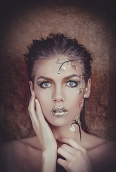 A striking portrait of a person with intricate gold and silver metallic makeup. The adornments resemble delicate leaves and branches, highlighting the eyes and lips. The background is a soft brown, enhancing the overall dramatic effect with a mix of natural and surreal elements.