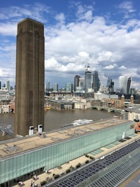 A large, industrial-looking tower stands prominently in the foreground, with a modern city skyline in the background featuring tall glass buildings. The river flows between the architectural structures, while further below, people are seen on a rooftop terrace. The sky is partly cloudy.