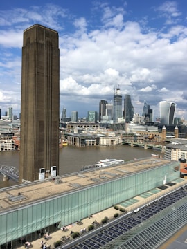 A large, industrial-looking tower stands prominently in the foreground, with a modern city skyline in the background featuring tall glass buildings. The river flows between the architectural structures, while further below, people are seen on a rooftop terrace. The sky is partly cloudy.