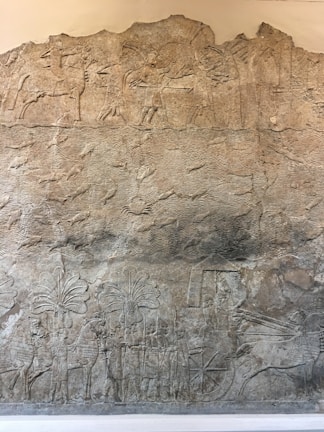 An ancient stone relief depicting a historical scene with figures in robes and helmets, horses, and a chariot. The top section shows marching soldiers, while the lower part features intricate carvings of palm trees and horses pulling a chariot. The relief is detailed with textures that indicate flowing water or desert terrain.
