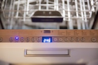 Close-up view of a dishwasher panel with various buttons and a digital display showing the time 2:45. The buttons include options like auto, eco, and different wash cycles. The background shows part of the dishwasher rack.