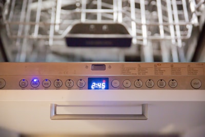 Close-up view of a dishwasher panel with various buttons and a digital display showing the time 2:45. The buttons include options like auto, eco, and different wash cycles. The background shows part of the dishwasher rack.