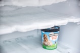 A container of Ben & Jerry's Cookie Dough ice cream is sitting inside a frosty freezer. The lid is black, and the container features a bright design with images of cookie dough and ice cream on a sky-blue background.