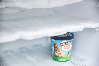 A container of Ben & Jerry's Cookie Dough ice cream is sitting inside a frosty freezer. The lid is black, and the container features a bright design with images of cookie dough and ice cream on a sky-blue background.