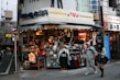 A street corner store selling various types of clothing, including jackets, shirts, and hats. The storefront is busy and colorful, with many items on display. Two people, a man and a woman, are walking past the store. Signage indicates a 24-hour operation, and several brand names like Levi's are visible.