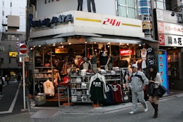 A street corner store selling various types of clothing, including jackets, shirts, and hats. The storefront is busy and colorful, with many items on display. Two people, a man and a woman, are walking past the store. Signage indicates a 24-hour operation, and several brand names like Levi's are visible.