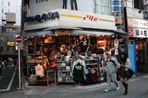 A street corner store selling various types of clothing, including jackets, shirts, and hats. The storefront is busy and colorful, with many items on display. Two people, a man and a woman, are walking past the store. Signage indicates a 24-hour operation, and several brand names like Levi's are visible.