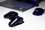 A black gaming console controller is placed on a light surface next to a sleek laptop and a gaming mouse with a blue LED light. The setting suggests a gaming or tech workspace with a focus on modern electronic devices.