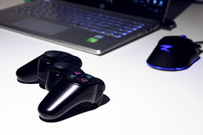 A black gaming console controller is placed on a light surface next to a sleek laptop and a gaming mouse with a blue LED light. The setting suggests a gaming or tech workspace with a focus on modern electronic devices.