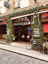 A quaint and inviting pub entrance adorned with lush greenery and signs promoting live music and comedy shows. The facade features vintage-style artwork and decorations, with a brick exterior typical of old town architecture. A bicycle rests casually against the wall, adding to the charming atmosphere. Warm lighting from inside emanates a cozy feel, while cobblestone streets in front suggest a historic setting.