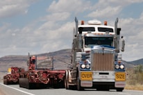 A large truck carrying an oversized load is driving on a road. The truck is predominantly white and blue with chrome detailing and has a yellow 'OVERSIZE' sign on the front. The road is bordered by mountainous terrain and a partly cloudy sky.