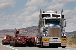 A large truck carrying an oversized load is driving on a road. The truck is predominantly white and blue with chrome detailing and has a yellow 'OVERSIZE' sign on the front. The road is bordered by mountainous terrain and a partly cloudy sky.