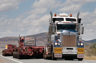 A large truck carrying an oversized load is driving on a road. The truck is predominantly white and blue with chrome detailing and has a yellow 'OVERSIZE' sign on the front. The road is bordered by mountainous terrain and a partly cloudy sky.