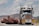 A large truck carrying an oversized load is driving on a road. The truck is predominantly white and blue with chrome detailing and has a yellow 'OVERSIZE' sign on the front. The road is bordered by mountainous terrain and a partly cloudy sky.