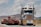 A large truck carrying an oversized load is driving on a road. The truck is predominantly white and blue with chrome detailing and has a yellow 'OVERSIZE' sign on the front. The road is bordered by mountainous terrain and a partly cloudy sky.
