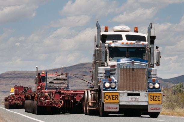 A large truck carrying an oversized load is driving on a road. The truck is predominantly white and blue with chrome detailing and has a yellow 'OVERSIZE' sign on the front. The road is bordered by mountainous terrain and a partly cloudy sky.