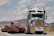 A large truck carrying an oversized load is driving on a road. The truck is predominantly white and blue with chrome detailing and has a yellow 'OVERSIZE' sign on the front. The road is bordered by mountainous terrain and a partly cloudy sky.