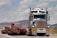 A large truck carrying an oversized load is driving on a road. The truck is predominantly white and blue with chrome detailing and has a yellow 'OVERSIZE' sign on the front. The road is bordered by mountainous terrain and a partly cloudy sky.