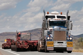 A large truck carrying an oversized load is driving on a road. The truck is predominantly white and blue with chrome detailing and has a yellow 'OVERSIZE' sign on the front. The road is bordered by mountainous terrain and a partly cloudy sky.