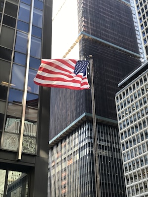 A United States flag waves in the foreground against a backdrop of tall, modern skyscrapers with reflective glass windows. The buildings have various architectural designs, including a grid-like pattern on the closest building.
