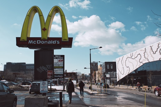 A McDonald's sign featuring a Canadian maple leaf stands prominently against a backdrop of an urban street. Several pedestrians walk along the sidewalk beside a line of parked cars. The sky is clear with a few fluffy clouds, and modern buildings with unique architectural designs are visible in the background.