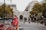 A man in a red jacket holds a microphone on a double-decker tour bus with empty red seats. The scene captures an urban setting with a white stone archway in the background, surrounded by historic buildings and lush trees with flags lined up along the sidewalk. The atmosphere indicates a city known for tourism.
