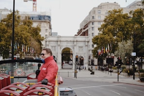 A man in a red jacket holds a microphone on a double-decker tour bus with empty red seats. The scene captures an urban setting with a white stone archway in the background, surrounded by historic buildings and lush trees with flags lined up along the sidewalk. The atmosphere indicates a city known for tourism.