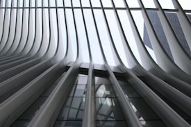 Curved, steel beams rise dramatically towards a glass ceiling, forming a striking architectural structure. The beams create a pattern of repeating lines and shadows, with glimpses of a skyscraper visible through the opening at the top.