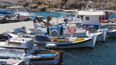 A busy marina with several small fishing boats docked along a pier. The boats have various equipment and supplies on them, such as nets and lifesavers. The water is a deep blue, contrasting with the white and blue color of the boats. In the background, rocky cliffs rise above the harbor.