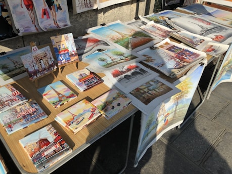 A collection of colorful paintings laid out on a table outdoors, depicting various scenes, including landmarks like the Eiffel Tower and vibrant street views. The paintings are displayed on a light wooden table under natural sunlight.