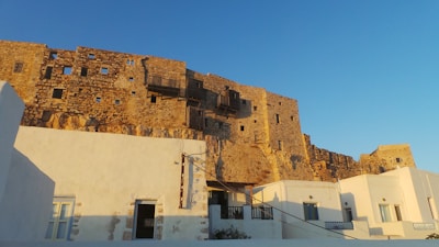 An ancient stone fortress with irregularly shaped windows stands atop a cliff, casting shadows in the warm evening light. Below, whitewashed buildings with blue doors and windows reflect the simple, traditional architecture of the region.