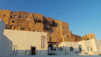 An ancient stone fortress with irregularly shaped windows stands atop a cliff, casting shadows in the warm evening light. Below, whitewashed buildings with blue doors and windows reflect the simple, traditional architecture of the region.