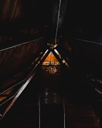 A dimly lit wooden attic space with a high, triangular ceiling supported by exposed beams. A vintage chandelier hangs at the center, casting a warm glow on the wooden surfaces around it.
