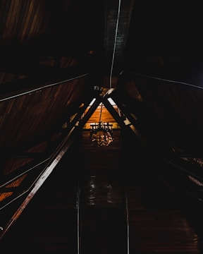 A dimly lit wooden attic space with a high, triangular ceiling supported by exposed beams. A vintage chandelier hangs at the center, casting a warm glow on the wooden surfaces around it.
