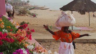A woman wearing a vibrant orange dress is balancing a large sack on her head while walking on a sandy beach. In the foreground, there are colorful flowers in shades of pink, red, and orange. The beach extends into the background with several traditional boats lined up along the shore. A thatched umbrella provides some shade, and several people are seen in the distance, contributing to a bustling seaside atmosphere.
