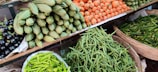 A vibrant assortment of fresh vegetables is displayed on shelves at a market. The arrangement includes carrots, green beans, spiny gourds, eggplants, and green chili peppers, all neatly organized in baskets or piles.