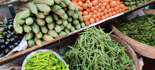 A vibrant assortment of fresh vegetables is displayed on shelves at a market. The arrangement includes carrots, green beans, spiny gourds, eggplants, and green chili peppers, all neatly organized in baskets or piles.