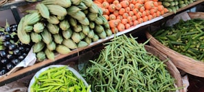 A vibrant assortment of fresh vegetables is displayed on shelves at a market. The arrangement includes carrots, green beans, spiny gourds, eggplants, and green chili peppers, all neatly organized in baskets or piles.