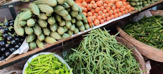 A vibrant assortment of fresh vegetables is displayed on shelves at a market. The arrangement includes carrots, green beans, spiny gourds, eggplants, and green chili peppers, all neatly organized in baskets or piles.