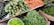 A vibrant assortment of fresh vegetables is displayed on shelves at a market. The arrangement includes carrots, green beans, spiny gourds, eggplants, and green chili peppers, all neatly organized in baskets or piles.