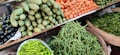 A vibrant assortment of fresh vegetables is displayed on shelves at a market. The arrangement includes carrots, green beans, spiny gourds, eggplants, and green chili peppers, all neatly organized in baskets or piles.