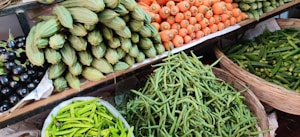 A vibrant assortment of fresh vegetables is displayed on shelves at a market. The arrangement includes carrots, green beans, spiny gourds, eggplants, and green chili peppers, all neatly organized in baskets or piles.