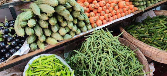 A vibrant assortment of fresh vegetables is displayed on shelves at a market. The arrangement includes carrots, green beans, spiny gourds, eggplants, and green chili peppers, all neatly organized in baskets or piles.