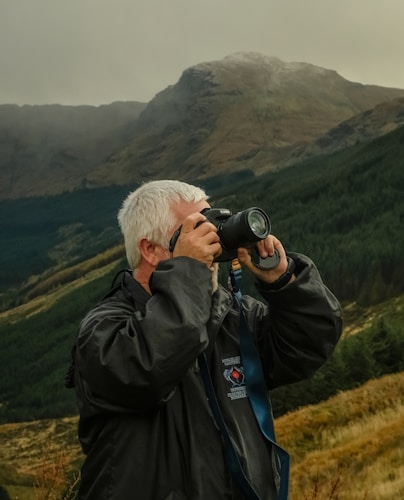 An individual with gray hair is using a camera in a scenic, mountainous landscape. They are wearing a dark jacket and appear to be focused on photographing the natural surroundings. The background features a misty mountain range and dense green forests.