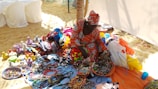A person is sitting on a sandy beach surrounded by vibrant, colorful handmade crafts and textiles. The crafts include woven baskets with lids and a variety of beaded jewelry items. The person is wearing a matching traditional outfit featuring bright patterns, adding to the burst of colors. In the background, there are white-covered chairs and a partial view of a beach tent.