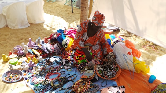 A person is sitting on a sandy beach surrounded by vibrant, colorful handmade crafts and textiles. The crafts include woven baskets with lids and a variety of beaded jewelry items. The person is wearing a matching traditional outfit featuring bright patterns, adding to the burst of colors. In the background, there are white-covered chairs and a partial view of a beach tent.