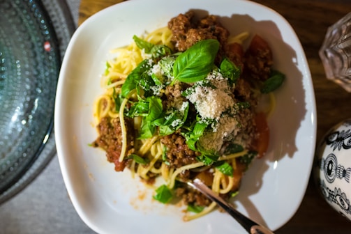 A bowl of spaghetti topped with a rich meat sauce, fresh basil leaves, and a generous sprinkle of grated parmesan cheese. The dish is set on a white plate with a fork resting on the side. A patterned cup and a textured glass plate are visible nearby.