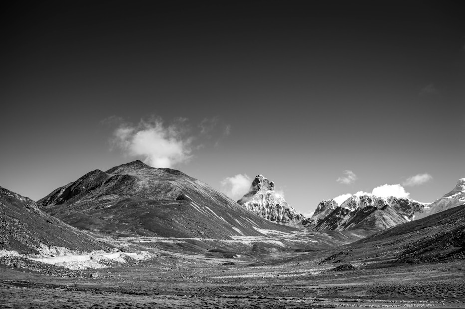 A black and white photograph of a mountainous landscape with rugged peaks and expansive valleys. Clouds gently float above the mountains, adding contrast to the otherwise barren terrain. The composition highlights the stark beauty of nature and the vastness of the scene.