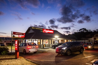 A fast-food restaurant with a prominent red sign is situated in the center, displaying 'red rooster'. Several parked and moving cars are present in the parking area and drive-thru lane, with visible headlights illuminating the scene. The sky exhibits a twilight hue, with scattered clouds adding to the evening ambiance.
