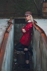 A person is standing between two rusted, concrete walls in an industrial setting, dressed in a red leather jacket and a plaid cap. The setting appears gritty and worn, with an overcast ambiance, creating a striking contrast with the bright attire.